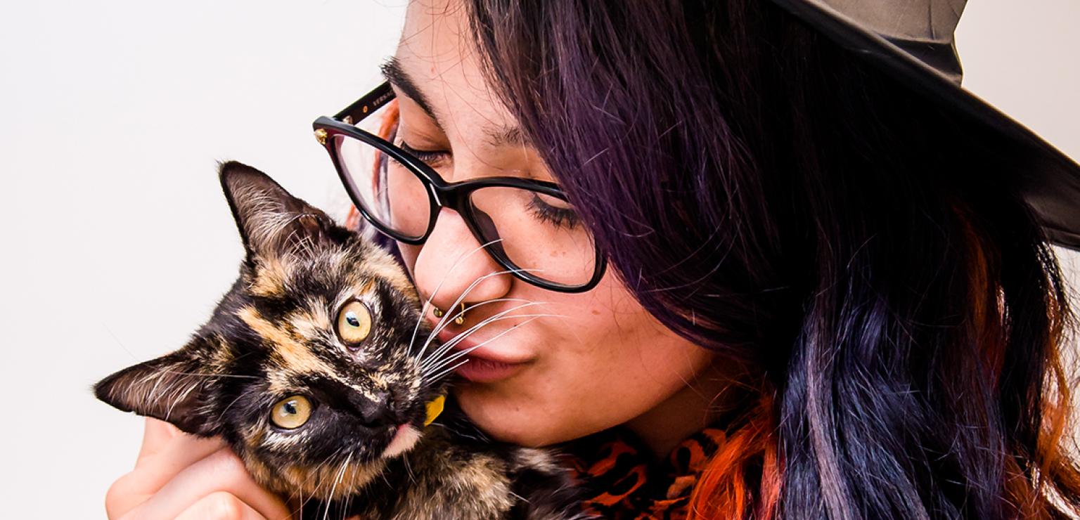 Person wearing a hat and colored hair kissing a tortoiseshell cat