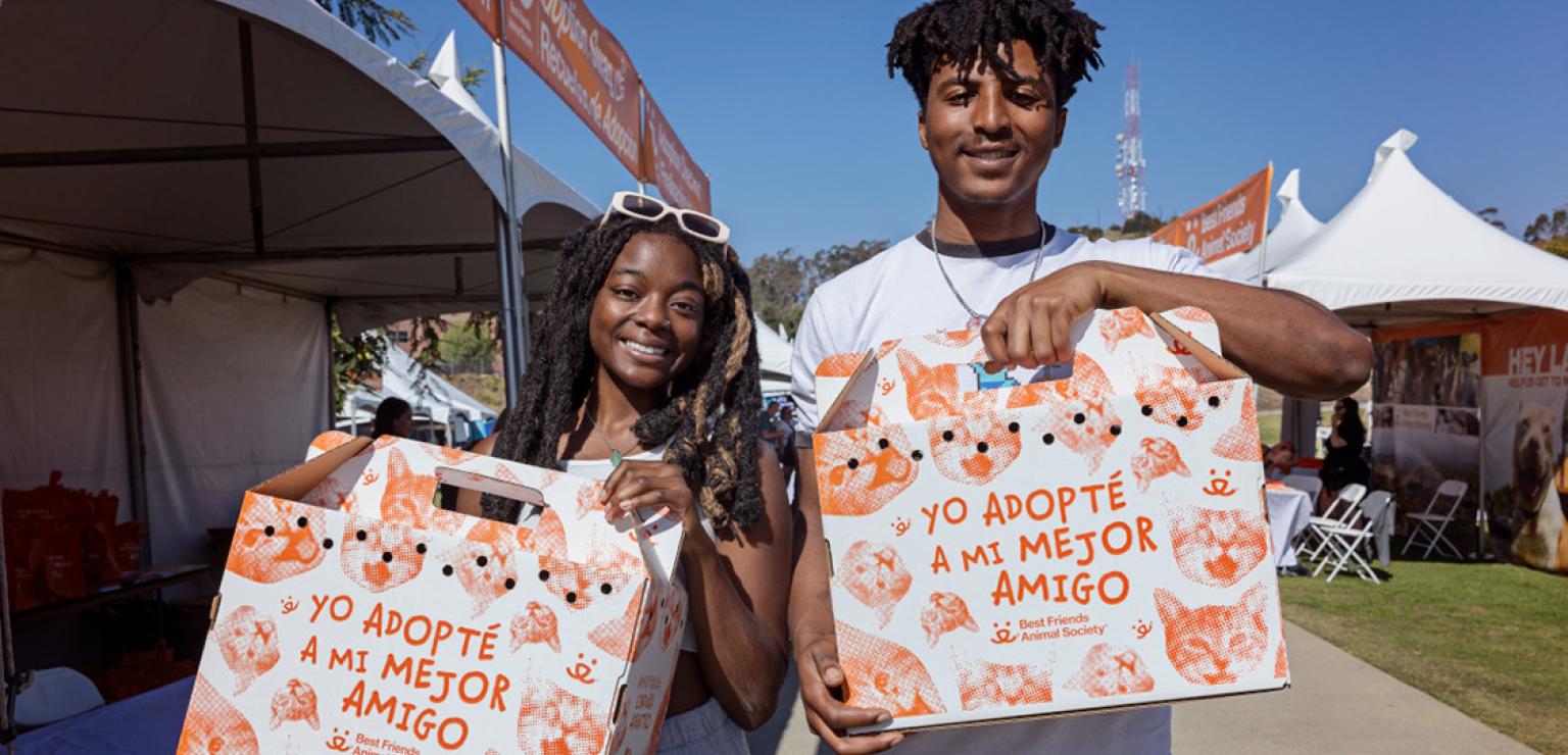 Two smiling people holding cardboard cat carriers with Spanish writing