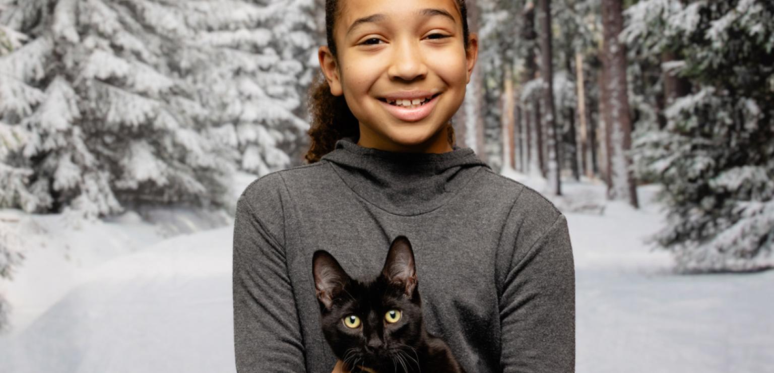 Smiling child holding a black kitten with a snowy evergreen backdrop
