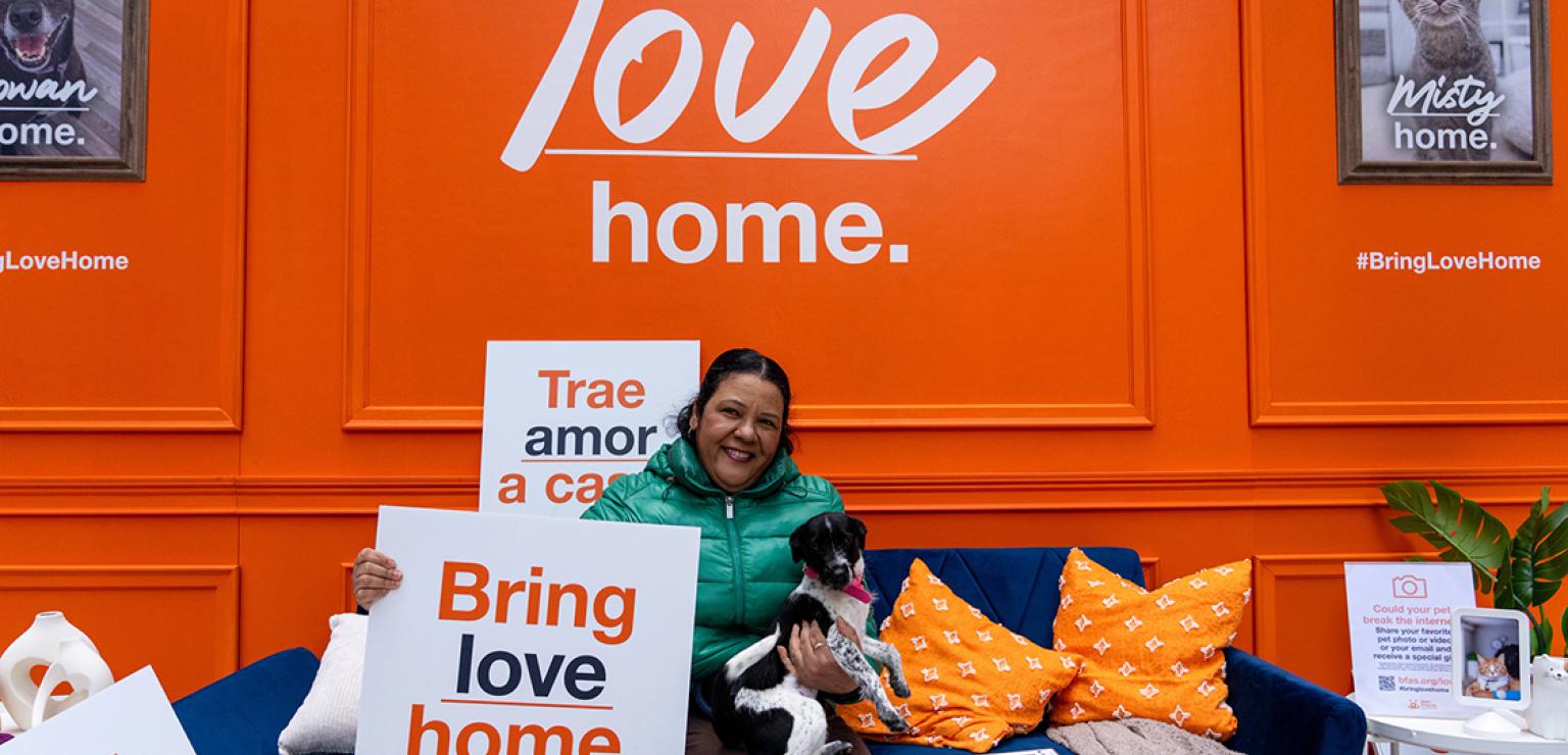 Person sitting on a couch at the New York Home campaign pop-up with a dog on her lap and a sign that says, "Bring love home."