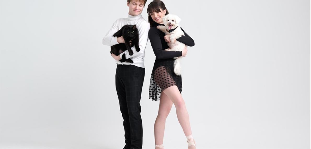 Two people in dance outfits, one holding a black cat and the other holding a white dog