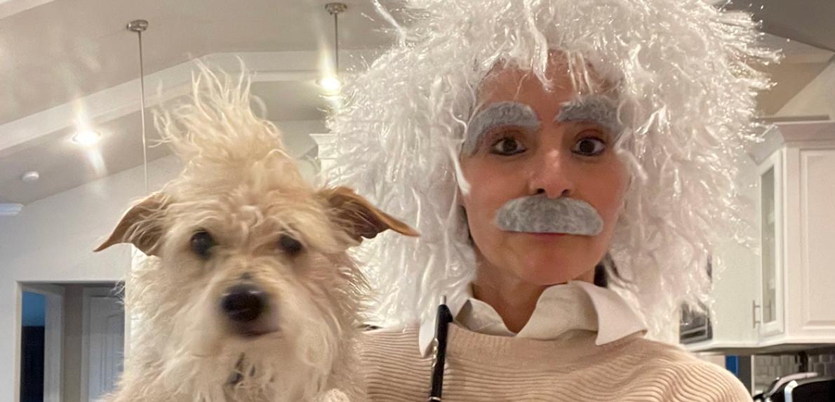 Person in a wig, mustache, and eyebrows to look like Albert Einstein next to fluffy white dog named Einstein
