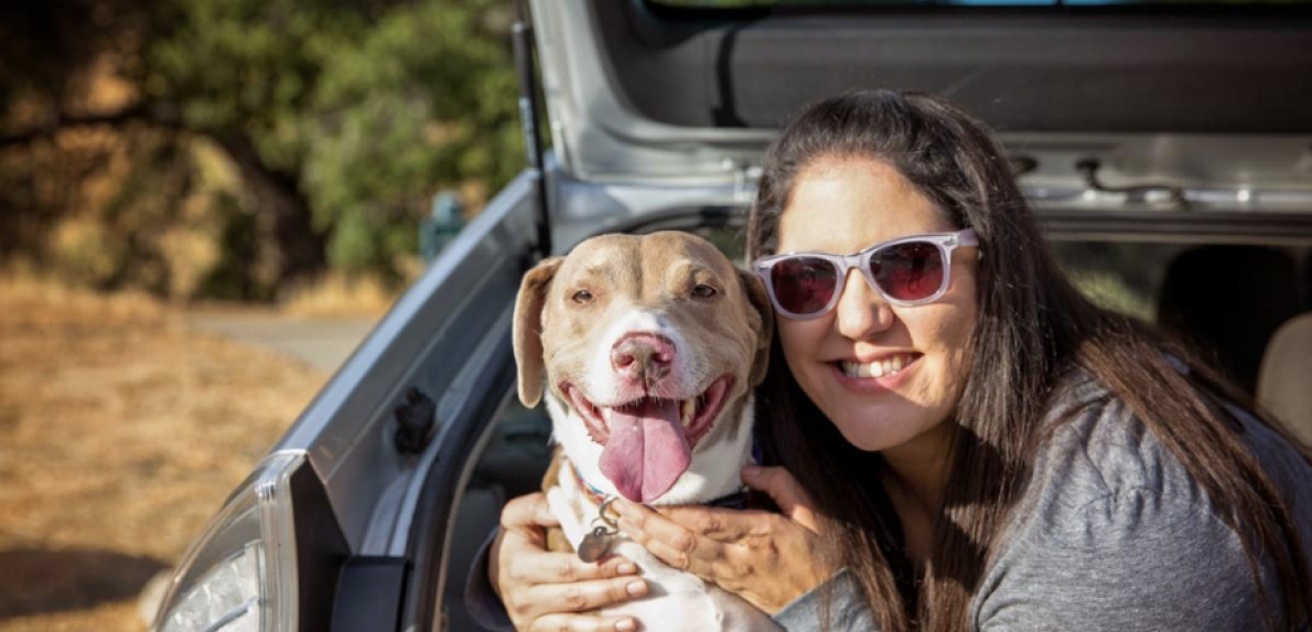Woman in sunglasses hugging a brown and white dog in the back of an open trunk