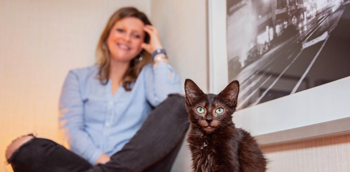 Black kitten in front of a smiling person who is sitting cross-legged next to a photo on the wall