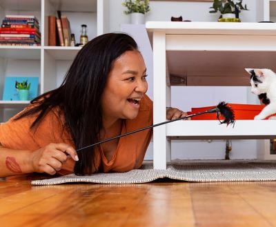 Smiling person lying on the floor using a wand toy to play with a black and white kitten