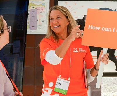 Person wearing a Best Friends orange volunteer T-shirt holding a sign that says, 'How can I help?' and speaking to a Best Friends National Conference attendee