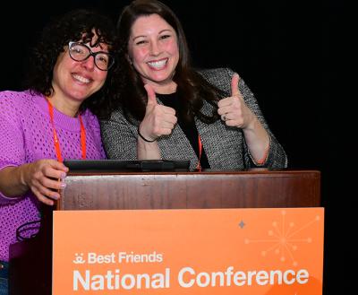 Two people behind a Best Friends National Conference podium, one giving a 'thumbs up' gesture with both hands