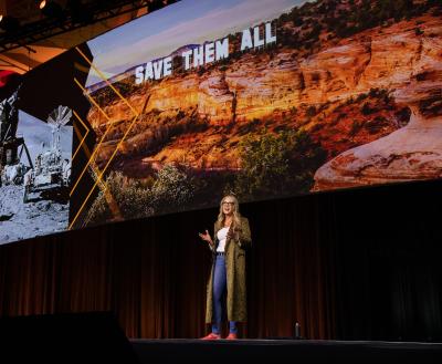 Julie Castle addressing the 2026 Best Friends National Conference attendees with giant slides of the moon landing and Angels Canyon behind her 