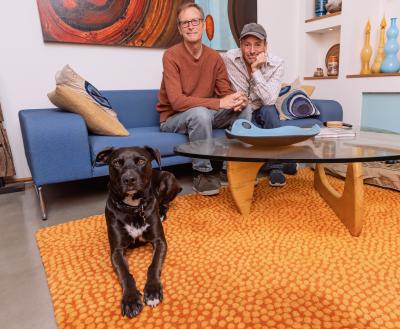 Two smiling people sitting on a couch behind a dog relaxing on the floor