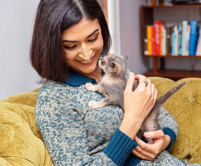 Smiling person cradling a kitten while sitting on a couch in front of a bookshelf