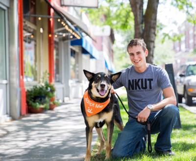 Smiling person wearing a NKUT T-shirt next to a dog wearing an Adopt Me bandana