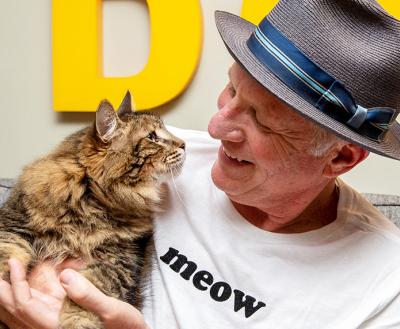 Smiling person wearing a hat and T-shirt that says 'meow' holding a brown tabby cat