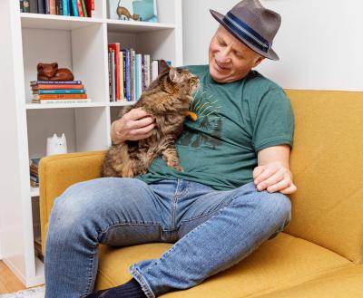 Person wearing a hat and sitting on a yellow couch with his hand on a brown tabby cat looking up at him