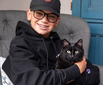 Child wearing a hat sitting in a chair snuggling with a black cat