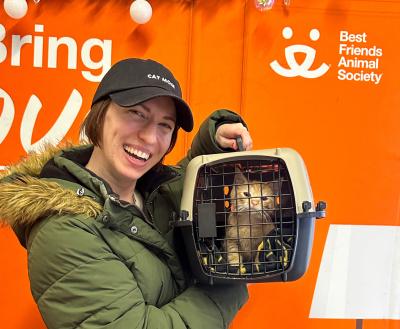Smiling person holding Joshua the cat in a carrier in front of a Best Friends backdrop