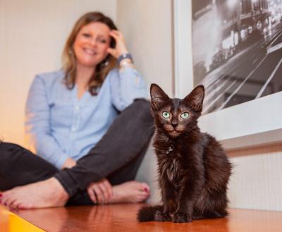 Black kitten in front of a smiling person who is sitting cross-legged next to a photo on the wall