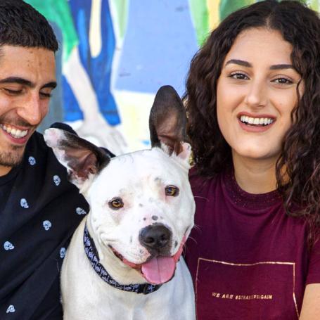 Two smiling people sitting with a dog outside in Los Angeles in front of a mural