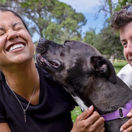Two people outside on grass by trees, smiling and interacting with a large dog