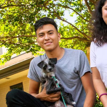 Two smiling people sitting in a yard with two small dogs