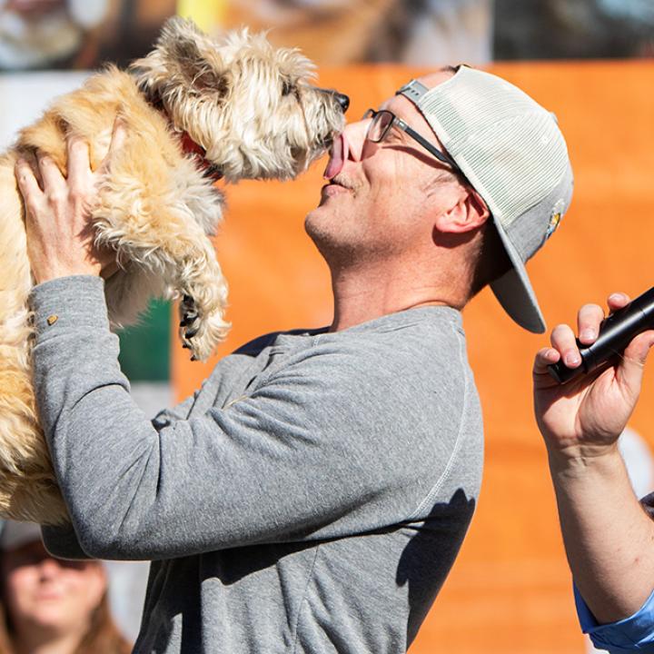 Small dog kissing the the nose of a person while another person wearing a Strut Your Mutt shirt holding a microphone watches