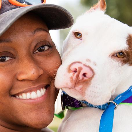 Smiling person with hat and sunglasses on top of head check-to-cheek with a pit-bull-type dog with cropped ears