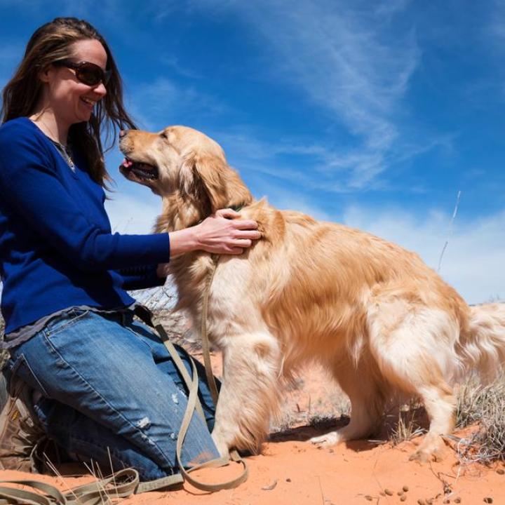Person kneeling with dog in red sand surrounded by desert brush and blue sky