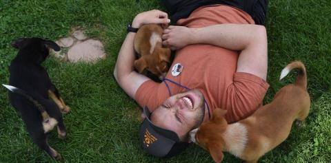 Guy in red shirt and baseball cap lying in the grass with puppy licking his face