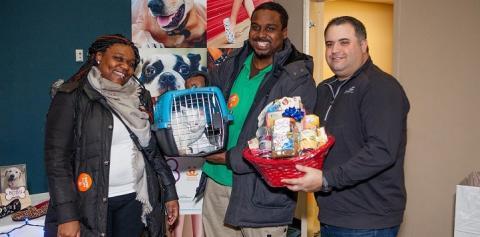 Woman standing next to man holding white cat in blue carrier next to a man holding a red gift basket