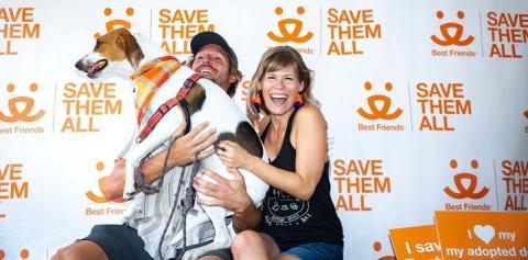 Man and woman holding tan and white hound dog in front of save them all background