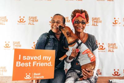 Dog wearing a gray T-shirt being held a person and kissing the face of another person who is holding a sign that says, 'I saved my Best Friend'