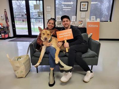 Bruno the dog lying on a couch with his two adopters, one who is holding a sign that says, I adopted my best friend