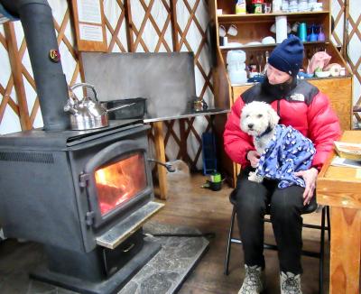 Kobe the little white dog in the lap of his adopter, both sitting by a wood burning stove