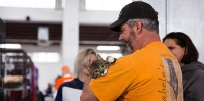 Man in black hat and yellow shirt cradling tabby cat looking at camera