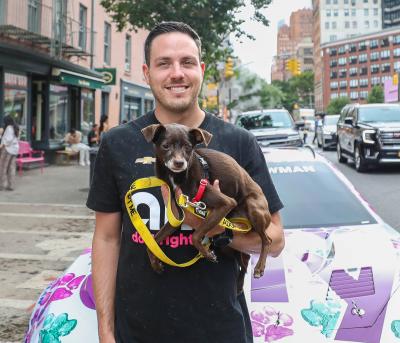 Alex Bowman of Ally Racing holding small brown dog in front of racing car parked in street
