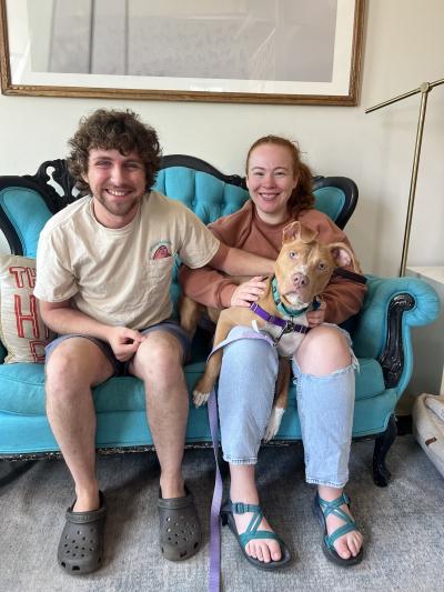 Two people smiling and sitting on a blue couch with a brown dog in one person's lap