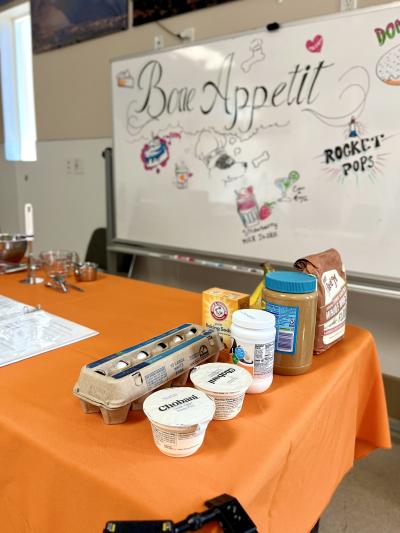 Table with an orange tablecloth and ingredients on it in front of a dry erase board that says, 'bone appétit'