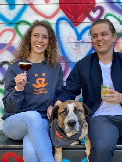 Two people, including one wearing a Best Friends logo sweatshirt and holding a glass of wine, next to Brutus the dog