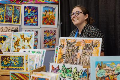 Smiling person at a vendor table displaying cat artwork