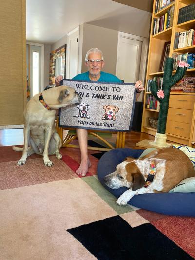 Cliff next to Yogi and Cliff the dogs holding up a welcome mat that says, Welcome to Yogi & Tank's van, pups on the run!