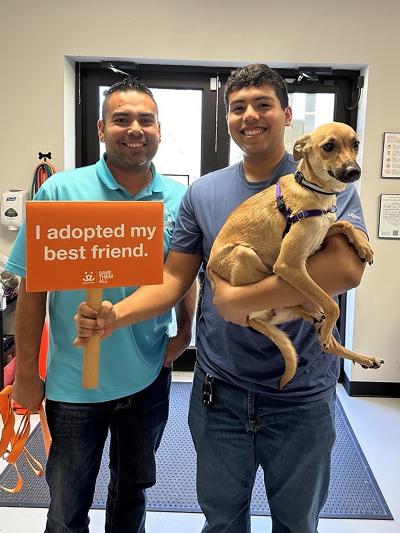 Kookie the dog being adopted, with one of the two people holding Kookie and a sign that says, 'I adopted my best friend'