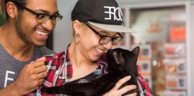 A person leaning over the shoulder of another person holding a black cat, all looking into each others eyes