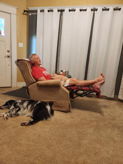 Jeff sitting in a recliner chair with Tator the dog in his lap and Bailey the dog lying down beside the chair