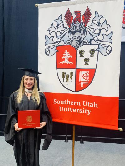 Jenny Franz holding her diploma at graduation next to a Southern Utah University sign