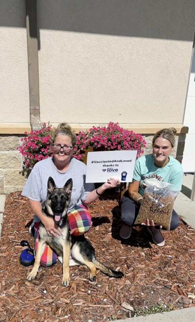 Shepherd-type dog with two people, one holding a bag full of dog kibble and one holding a sign that says, 'vaccinated and loved thanks to Petco Love'