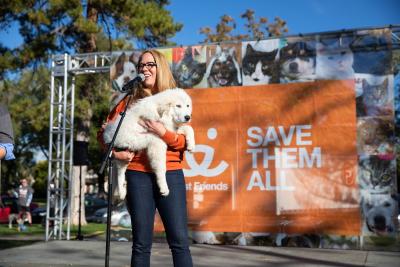 Julie Castle at a microphone holding a white dog with a Save Them All Best Friends banner behind her
