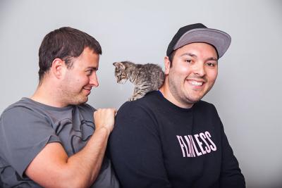 Photo of Meeko the kitten on the shoulder of one of his adopters while the other adopter smiles at him