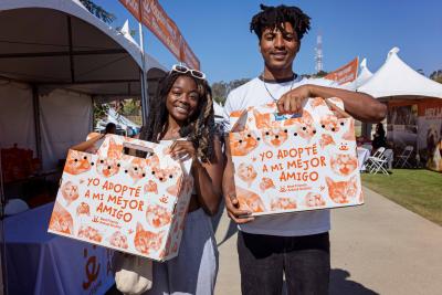 Two smiling people, each holding a cardboard cat carrier printed with Spanish text and cat graphics on them