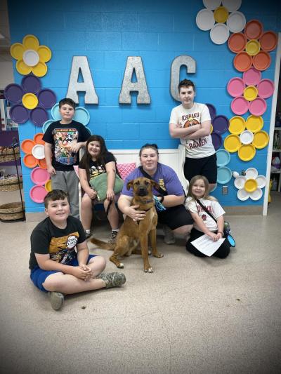 Brody the dog surrounded by people with a blue wall with flowers and AAC behind them