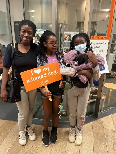 Family of three adopters with one holding a black and white puppy in a lavender blanket and another holding a "I heart my adopted dog" sign at the Best Friends Lifesaving Center in New York City