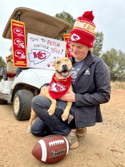 Norberta, one of the dogs who inspired the cleats, wearing a Kansas City Chiefs bandana with a football and signage and a person who is wearing a team hat